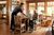 A server delivers food to a smiling older couple seated at a wooden table in a warmly lit dining area at Custer State Park. The room features rustic decor and large windows with views of greenery outside.