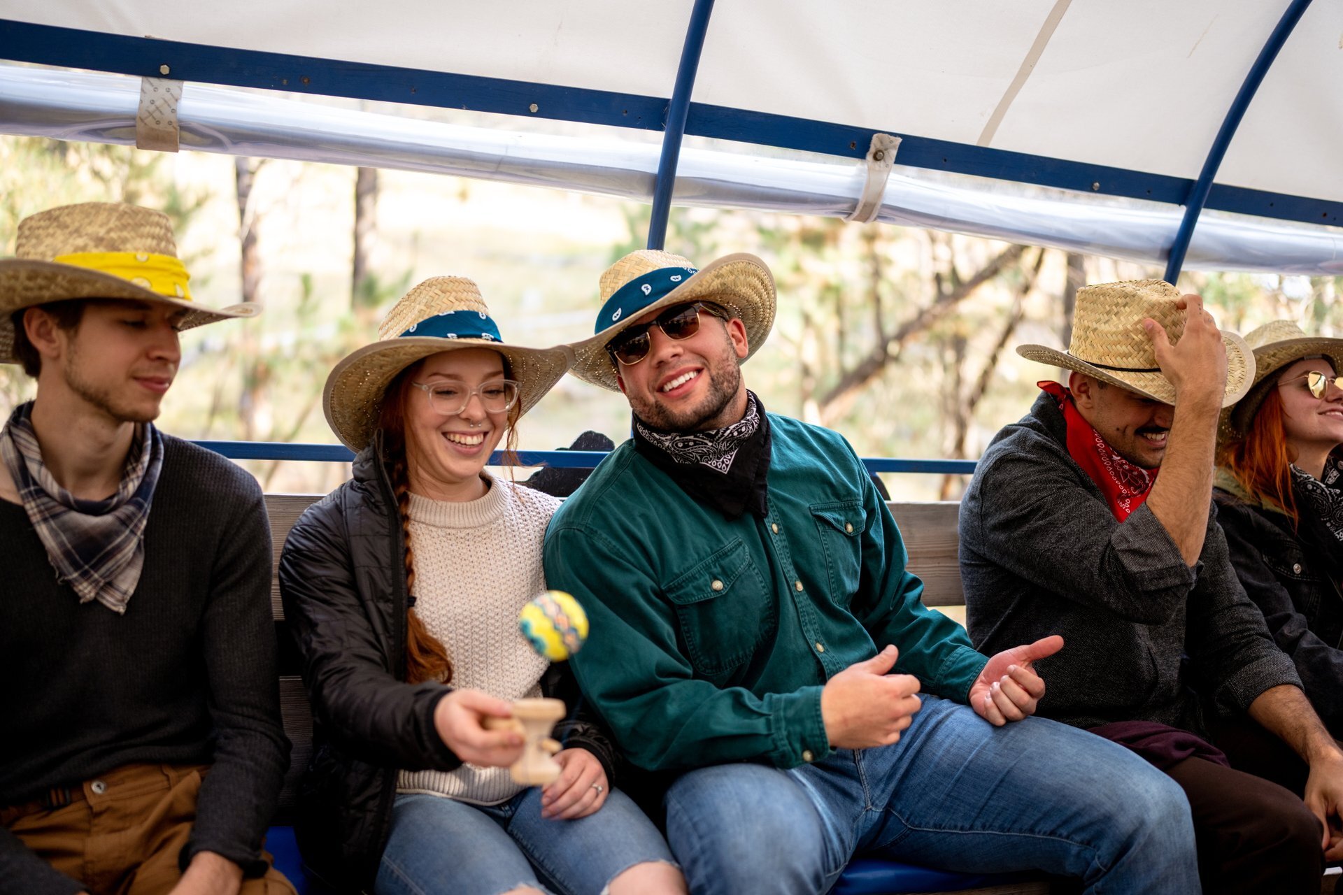 A group of smiling young adults in cowboy hats enjoy a covered wagon ride at Custer State Park. One woman plays with a toy while others laugh and relax, dressed in bandanas and Western-style outfits.
