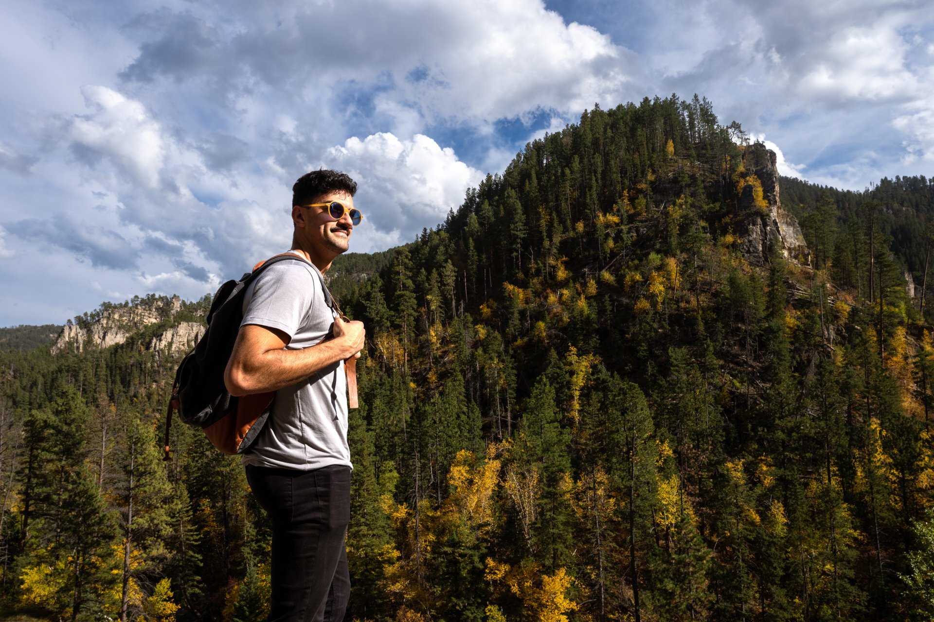 Smiling man wearing sunglasses and a backpack stands on a scenic overlook, surrounded by forested hills and rocky cliffs in Custer State Park. The sky is partly cloudy with sun shining through.