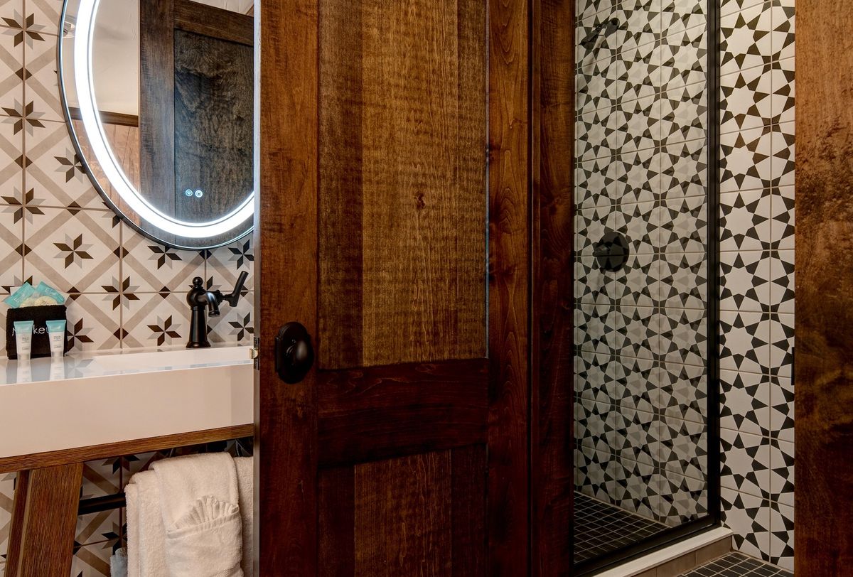 Bathroom with dark wood finishes, patterned tile, and a circular lighted mirror above a modern vanity.