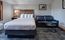 Modern hotel room with a king-sized bed, five white pillows, a dark leather loveseat, and two round nesting tables. The space features rustic wood accents, gray carpet, and a framed bison photo on the wall.
