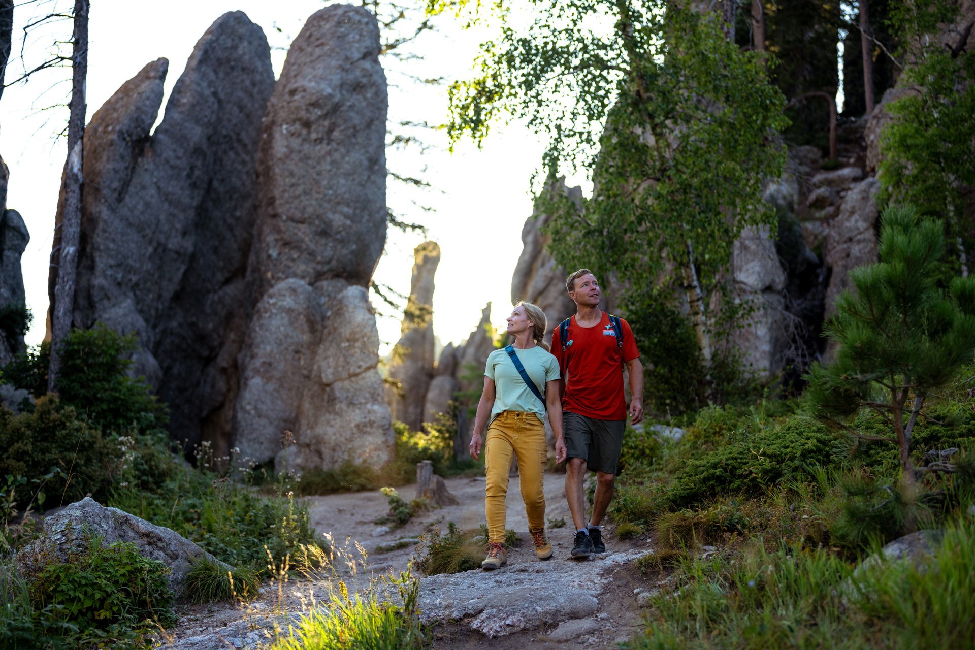 A couple hikes through a forest trail surrounded by towering granite spires in Custer State Park. They hold hands and look upward, bathed in soft sunlight filtering through the trees.