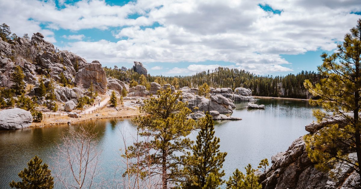 A small lake, surrounded by a rocky shoreline and hills. Besides the large boulders, pine trees fill the hills that surround the lake. The sky is blue with scattered white, puffy clouds. A small gravel path runs along the shoreline on the other side of the lake from where the photo was taken.