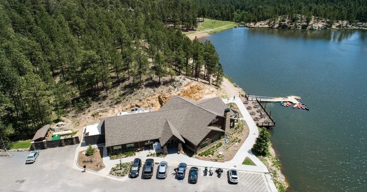 An aerial image of Legion Lake Lodge. It's on the shore, with a dock and small boats floating in the water, and pine trees lining the hills behind it.