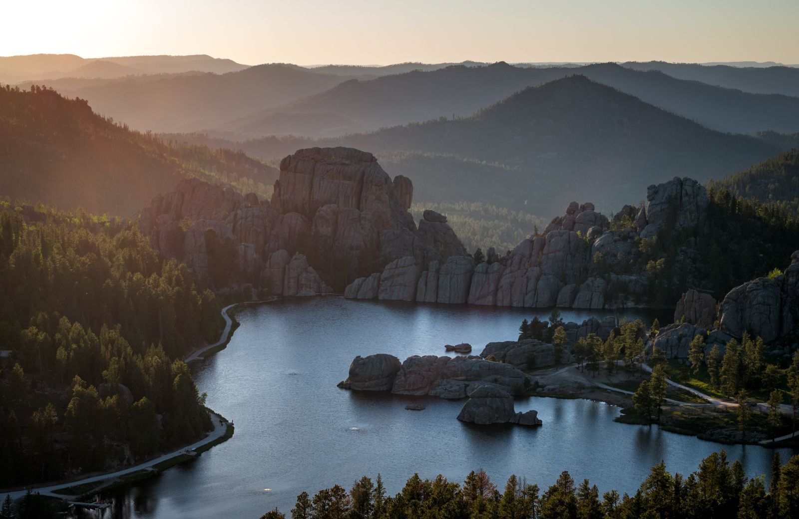 Aerial view of Sylvan Lake in Custer State Park at sunset, with dramatic rock formations rising from the water and tree-covered hills fading into the distance. Warm light bathes the landscape in a golden glow.