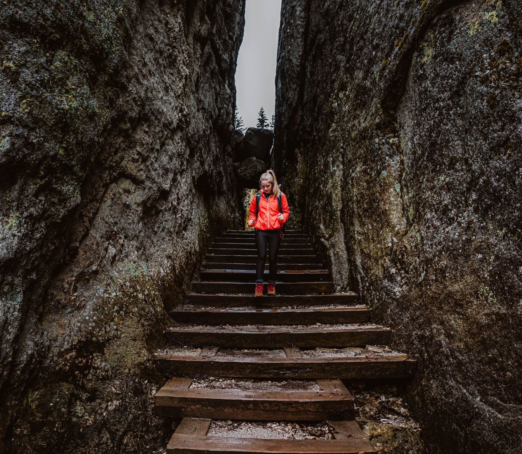 A woman in a bright orange jacket descends wooden steps through a narrow rock passage in Custer State Park. Towering stone walls rise on both sides, creating a dramatic and rugged landscape.